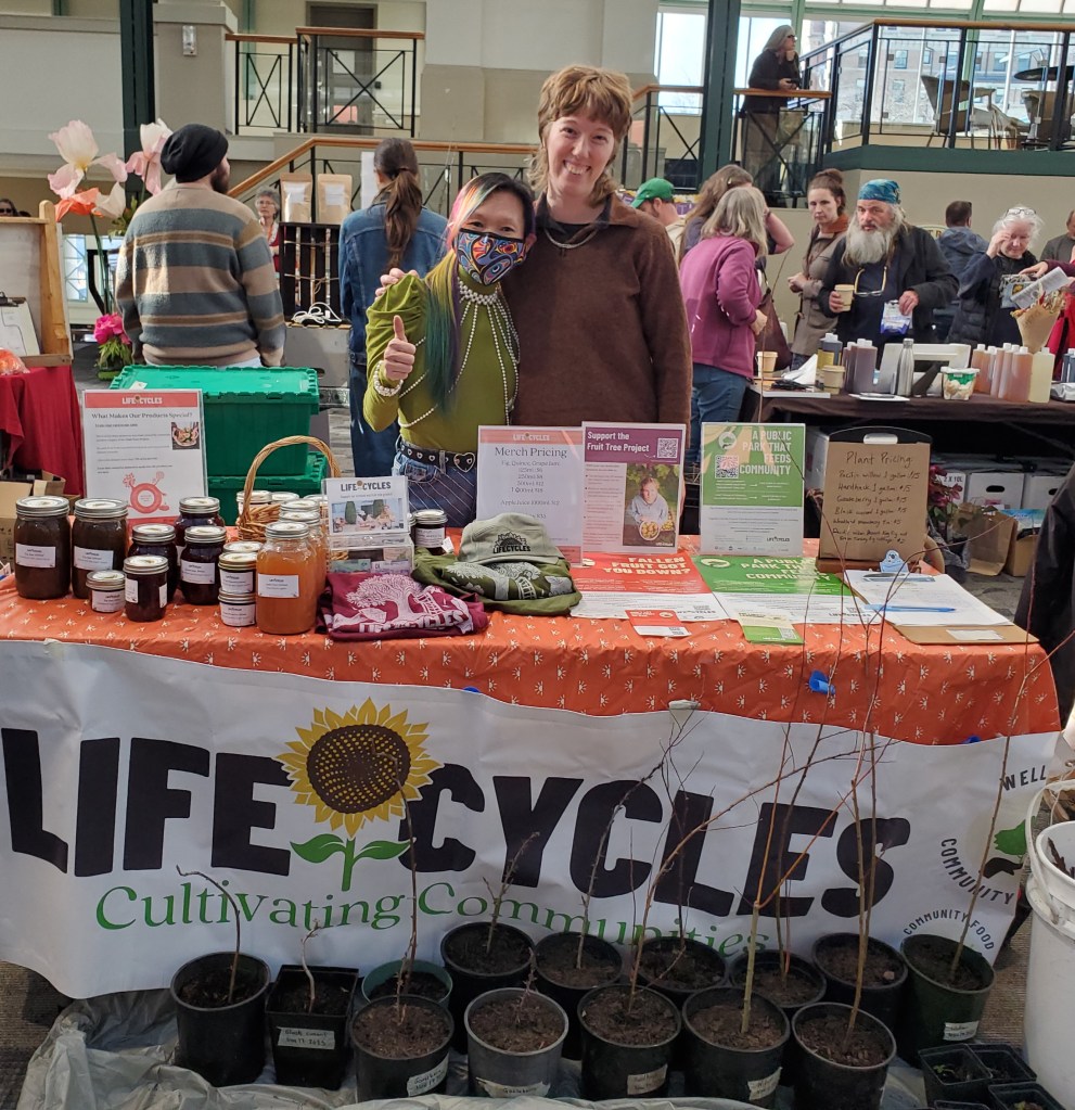 Two women smiling behind a "Life Cycles" market table showcasing jams, merchandise, and potted saplings, with signage promoting community fruit tree projects and plant pricing, surrounded by other vendors and shoppers in an indoor event space.